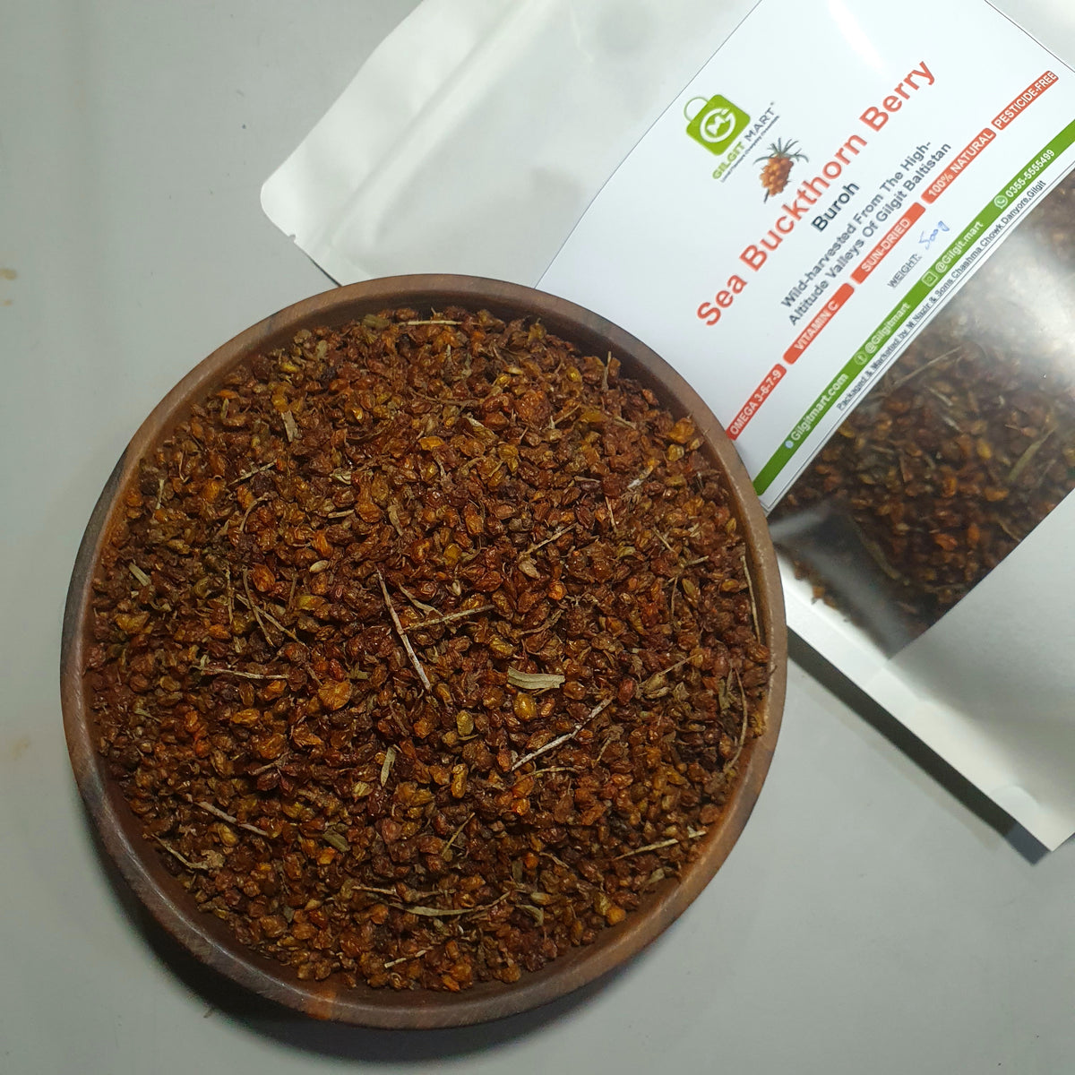 A wooden bowl filled with dried red-brown sea buckthorn berries sits in front of a white stand-up pouch labeled 'Sea Buckthorn Berry' on a light gray surface.
